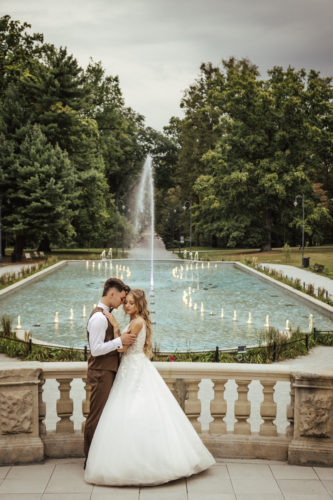 Przytuleni czołami Patrycja i Bartek na sesji poślubnej w parku z fontanną